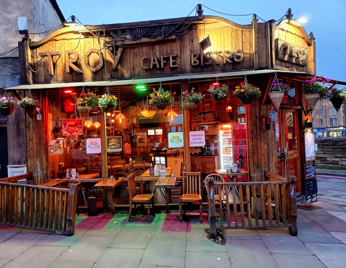 Troy Cafe-Bistro's carved timber shopfront at dusk, glowing amber with hanging flower baskets, Dalry Road Edinburgh